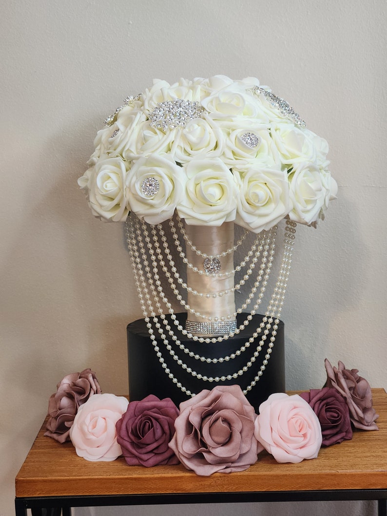 May include: A white bridal bouquet with pearl accents and rhinestone embellishments. The bouquet is surrounded by smaller blush and burgundy roses.