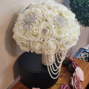 May include: A white bridal bouquet made of artificial roses with pearl and rhinestone embellishments. The bouquet is sitting on a black pedestal and surrounded by smaller artificial roses in shades of pink.