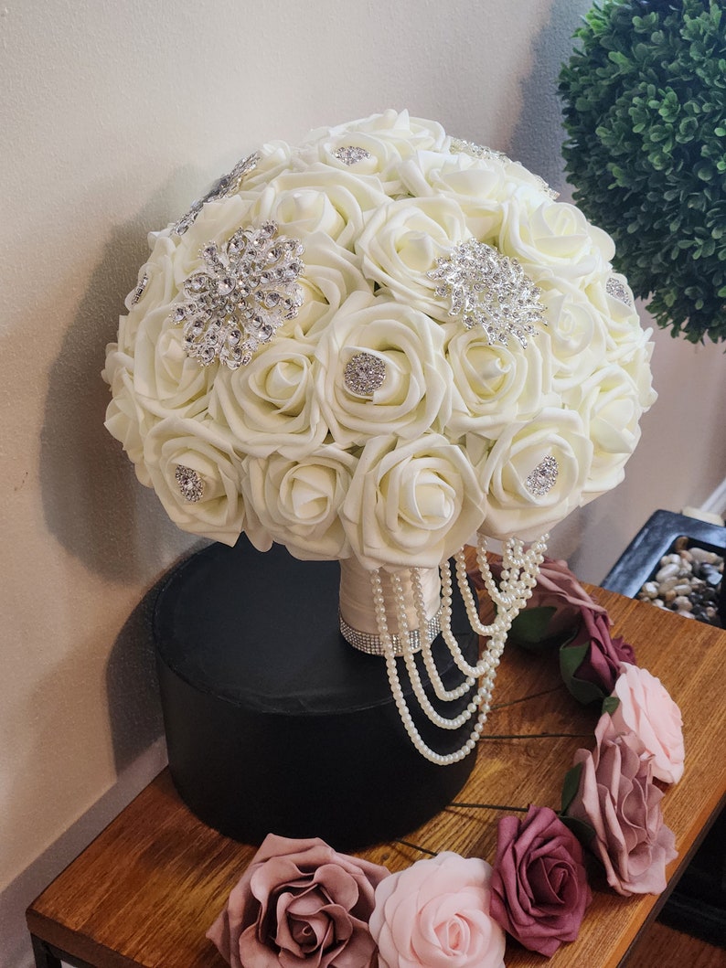 May include: A white bridal bouquet made of artificial roses with pearl and rhinestone embellishments. The bouquet is sitting on a black pedestal and surrounded by smaller artificial roses in shades of pink.