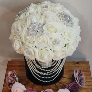 May include: A white bridal bouquet made of artificial roses with pearl and rhinestone embellishments. The bouquet is in a black vase with a pearl garland. The bouquet is surrounded by smaller pink and burgundy roses.