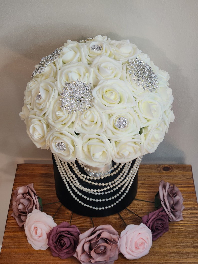 May include: A white bridal bouquet made of artificial roses with pearl and rhinestone embellishments. The bouquet is in a black vase with a pearl garland. The bouquet is surrounded by smaller pink and burgundy roses.