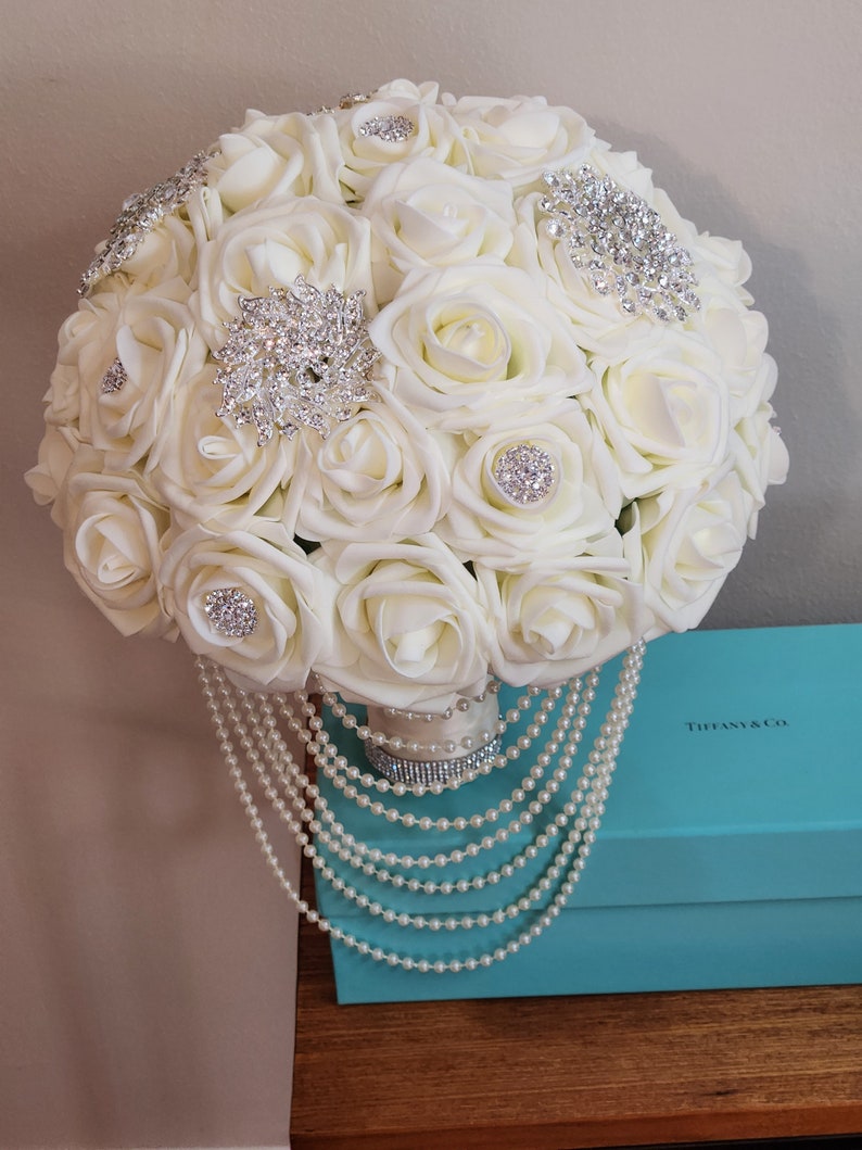May include: A white bridal bouquet made of artificial roses with pearl accents and rhinestone embellishments. The bouquet is sitting on top of a Tiffany & Co. blue box.