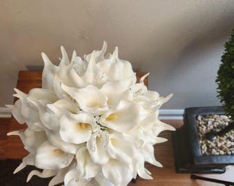May include: A bouquet of white calla lilies with yellow centers. The flowers are arranged in a dense, rounded shape, with each petal gracefully curved. Clear, decorative accents are interspersed throughout the arrangement, adding a touch of sparkle.