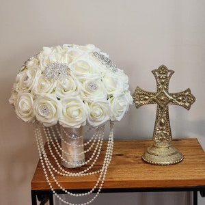May include: A white bridal bouquet with pearl accents and rhinestone embellishments. The bouquet is on a wooden table next to a gold cross.