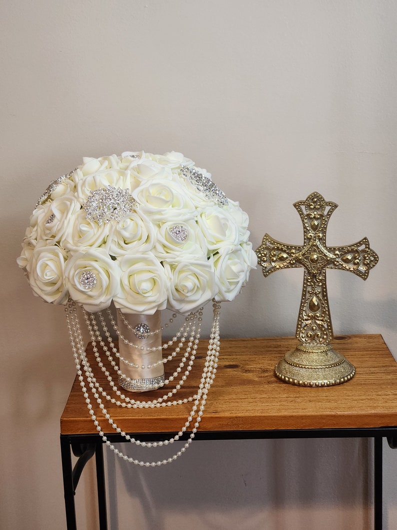 May include: A white bridal bouquet with pearl accents and rhinestone embellishments. The bouquet is on a wooden table next to a gold cross.