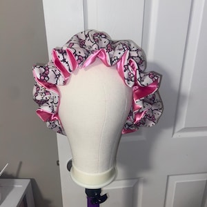 May include: A white satin bonnet with a repeating pattern of pink cartoon characters. The bonnet has a pink satin trim and is displayed on a white mannequin head. The bonnet is designed to protect hair.