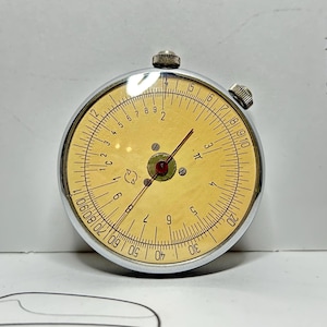 以下が含まれることがあります： シルバーの金属ケースとクリーム色の文字盤を備えた、ヴィンテージの円形計算尺。 文字盤には黒と赤のマーキング、数字、赤い針があります。 上部と側面に2つの小さなノブが見えます。