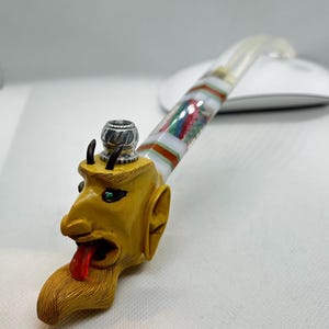 Puede incluir: Una pipa de cerámica amarilla y blanca con un diseño de cabeza de diablo. La pipa tiene una cazoleta de plata y una boquilla larga y delgada.