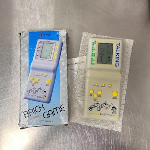 Op de afbeelding: Een beige draagbare elektronische spelcomputer met het label Brick Game 96 in 1 E-23 Plus Mark II met gele knoppen en een digitaal scherm, vergezeld van de originele blauw-witte kartonnen verpakking en een gedrukt instructieblad.