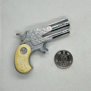 Op de afbeelding: Een zilverkleurig, gegraveerd, dubbelloops pistool met een witte kunststof handgreep. Het pistool ligt naast een zilveren munt met de tekst "10 ГРИВЕНЬ" erop.