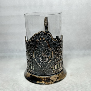 Può includere: Porta bicchiere in argento con un design ornato e un bicchiere di vetro trasparente. Il porta bicchiere presenta un'immagine dettagliata di un edificio e motivi floreali.