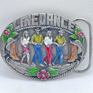以下が含まれることがあります： 上部に「LINEDANCE」の文字が入った、銀色の楕円形のベルトバックル。バックルには、カウボーイブーツと帽子を身に着けた4人のラインダンスをする人々のレリーフが施されています。デザインには、花のモチーフと羽のアクセントが含まれています。