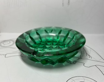 Könnte beinhalten: Ein runder, smaragdgrüner Aschenbecher mit facettiertem Design. Der Glasaschenbecher hat eine kreisförmige Form mit einem leicht erhöhten Rand und einer Reihe vertikaler Rillen. Der Aschenbecher liegt auf einer weißen Oberfläche.