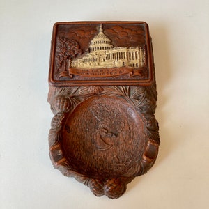 Op de afbeelding: Een houten asbak met een gegraveerd beeld van het US Capitol gebouw in Washington D.C. erop. De asbak is bruin en heeft een gegraveerd dennenappelontwerp rond de rand.