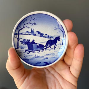 May include: A small, round, blue and white ceramic plate with a winter scene. The scene depicts a horse-drawn sleigh with two people riding in it, traveling through a snowy landscape with a small village in the background.