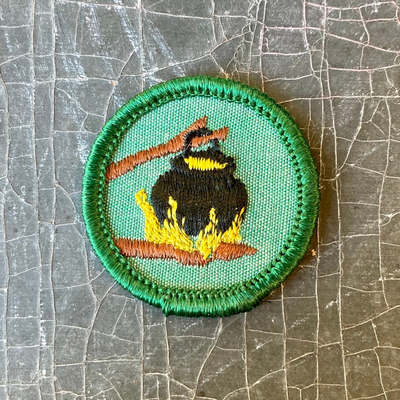 Puede incluir: Un parche bordado verde y marr&oacute;n con una olla negra sobre un fuego. El parche es una insignia de Girl Scout.