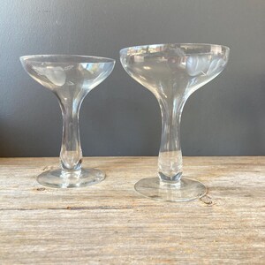 May include: Two clear champagne coupe glasses with etched floral designs. The glasses have a wide, shallow bowl and a long, slender stem. The etched design includes a heart and leaf pattern. The glasses are displayed on a wooden surface.
