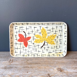 May include: A white metal tray with a black grid pattern and two painted leaves. One leaf is yellow and the other is orange.