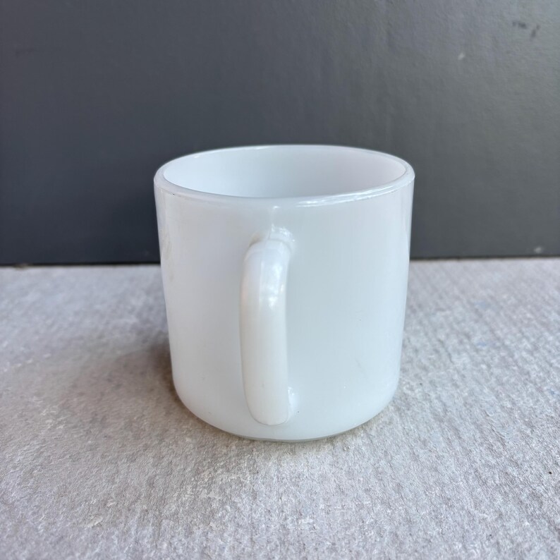 May include: A white, opaque mug with a rounded body and a curved handle. The mug is made of a solid, smooth material and is set against a neutral background. The handle is attached to the side of the mug.