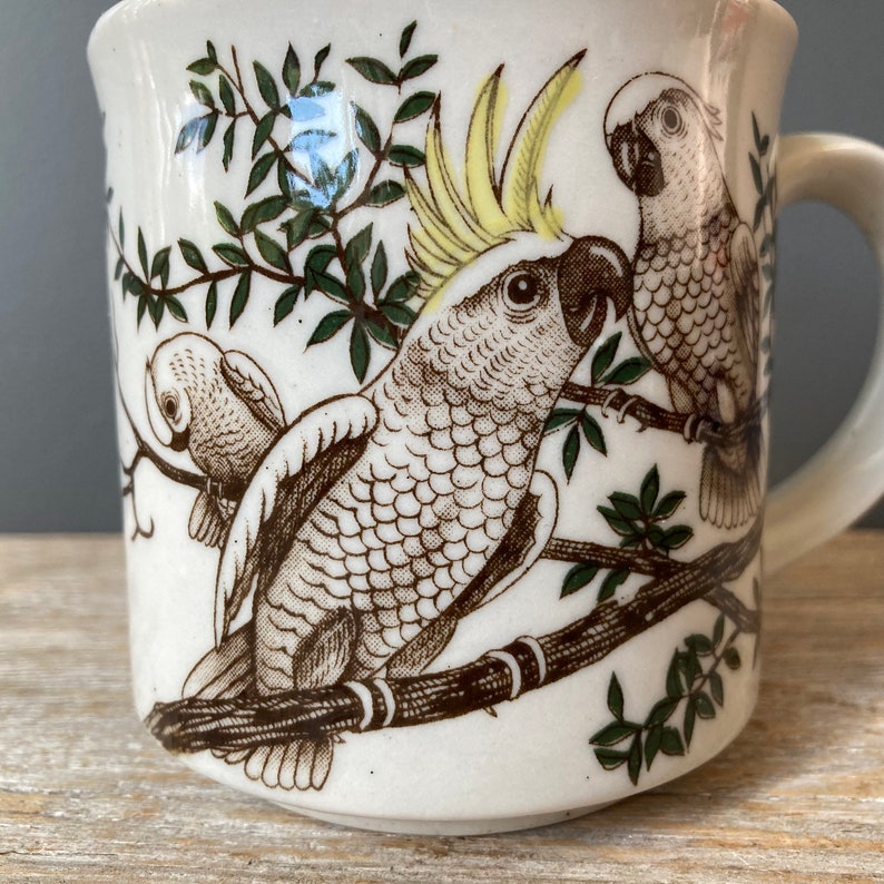 May include: White ceramic mug with a brown and green design of three cockatoos perched on branches. The cockatoos have yellow crests and brown and white feathers.