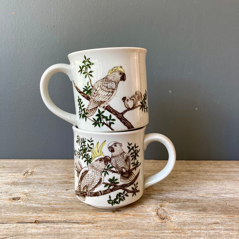 May include: Two white ceramic mugs with brown and green floral designs featuring white cockatoos. The mugs are stacked on top of each other.