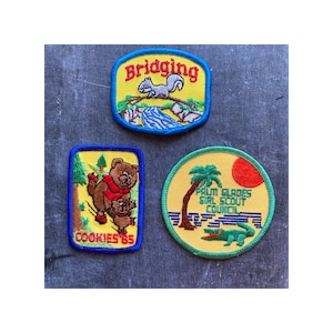May include: Three embroidered patches with a yellow background. The top patch is blue and says "Bridging" with a squirrel on a log. The left patch is blue and says "Cookies '85" with a bear and a cub. The right patch is green and says "Palm Glades Girl Scout Council" with a palm tree and an alligator.