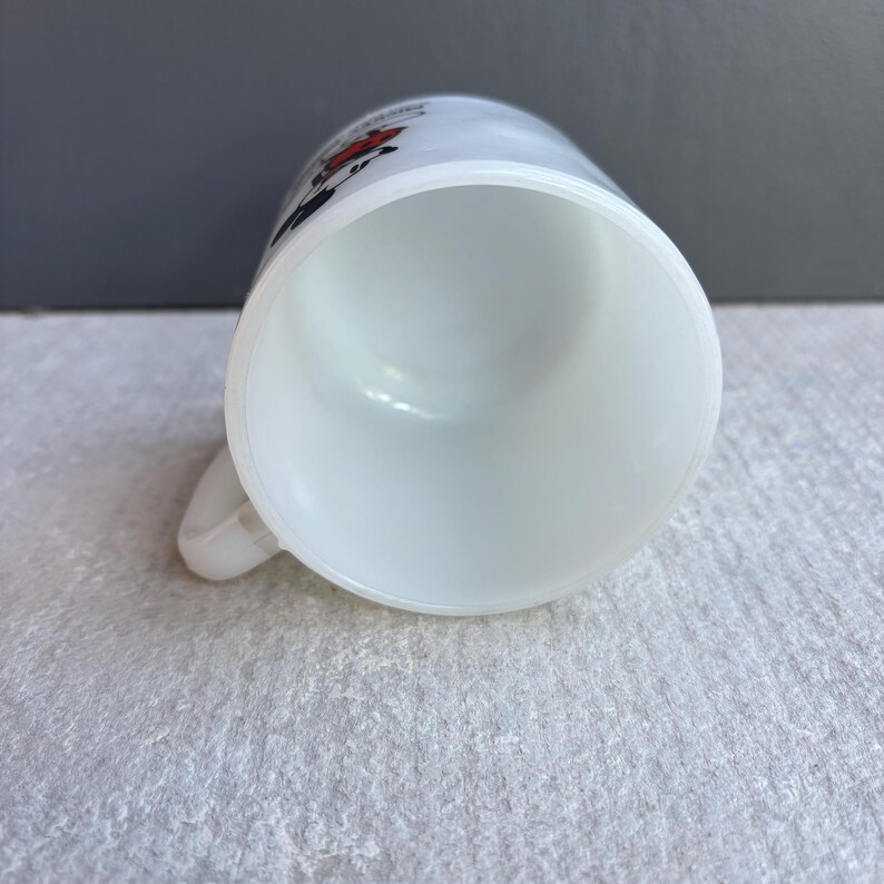 May include: White ceramic mug with a handle. The mug has a slightly flared rim and a smooth, glossy finish. The top of the mug has a design with red and black accents. The mug is empty and clean.