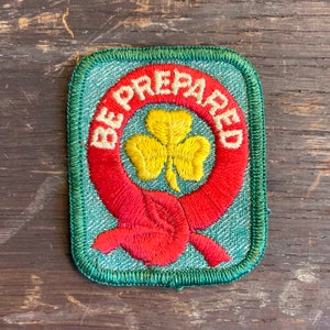 May include: A rectangular patch with a green border and a light blue background. The patch features a red circle with the words "BE PREPARED" in white. Inside the circle is a yellow shamrock.