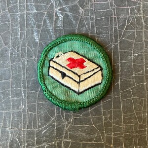 Puede incluir: Un parche bordado verde y blanco con una cruz roja sobre un botiqu&iacute;n de primeros auxilios blanco. El parche est&aacute; rodeado por un borde verde.