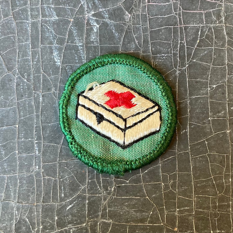Puede incluir: Un parche bordado verde y blanco con una cruz roja sobre un botiqu&iacute;n de primeros auxilios blanco. El parche est&aacute; rodeado por un borde verde.
