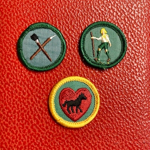 May include: Three circular embroidered patches with green borders. One shows crossed paintbrushes, another a person with a staff, and the third a black horse inside a red heart, with a yellow border.