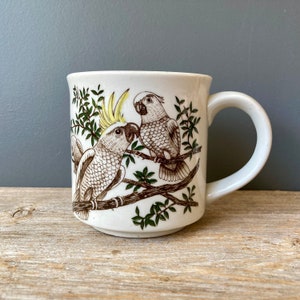 May include: White ceramic mug with a brown and green design of two cockatoos perched on branches. The mug has a white handle.