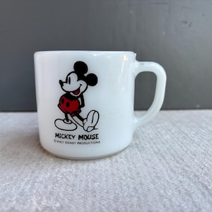May include: White ceramic mug featuring Mickey Mouse. The iconic character is depicted in black, red, and white. The mug has a curved handle and the text "MICKEY MOUSE" and "WALT DISNEY PRODUCTIONS" printed on the base.