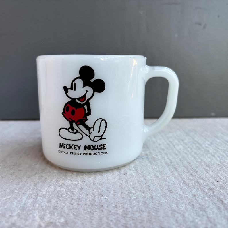 May include: White ceramic mug featuring Mickey Mouse. The iconic character is depicted in black, red, and white. The mug has a curved handle and the text "MICKEY MOUSE" and "WALT DISNEY PRODUCTIONS" printed on the base.