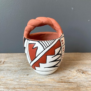 May include: Handmade ceramic basket with a twisted handle. The basket features a white base with geometric designs in red and black. The handle is a reddish-brown color. The basket is sitting on a wooden surface.