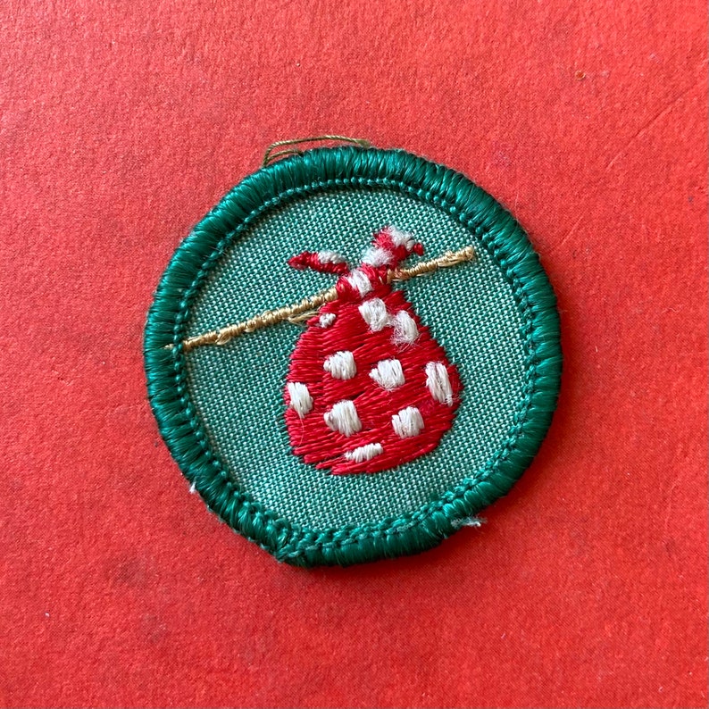 May include: A green and white embroidered Girl Scout badge featuring a red and white polka dot bag hanging from a gold string.