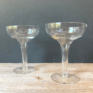 Könnte beinhalten: Zwei transparente Champagner-Coupe-Gläser. Jedes Glas hat eine breite, flache Schale auf einem langen Stiel, der sich zur Basis hin ausdehnt. Die Gläser bestehen aus transparentem Glas und reflektieren Licht und Umgebung.