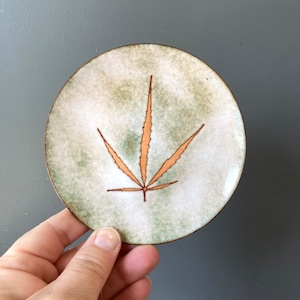 May include: A small, round, ceramic plate with a green and white glaze. The plate features a brown leaf design in the centre.