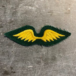 May include: A green fabric patch with yellow embroidered wings. The patch is slightly frayed around the edges.