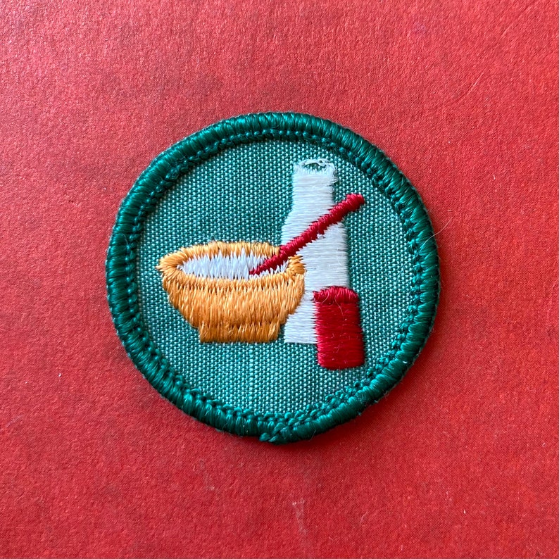 May include: A green and white embroidered Girl Scout badge with a bowl, spoon, and bottle. The badge is for the "Cooking" merit badge.