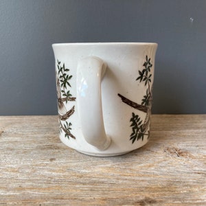 May include: White ceramic mug with a brown and green bird design. The mug has a white handle.