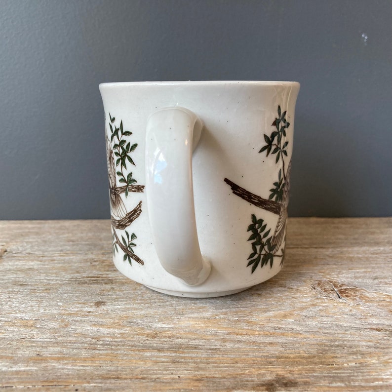 May include: White ceramic mug with a brown and green bird design. The mug has a white handle.