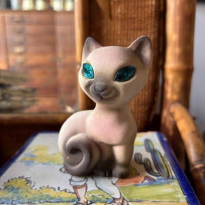 May include: A ceramic Siamese cat figurine with a light beige body, darker brown ears, face, and tail, and bright teal faceted glass eyes. The cat is in a seated position with its tail curled around its side, featuring a smooth matte finish.