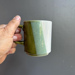 May include: A ceramic mug with a unique color block design. The mug features a combination of olive green, speckled seafoam green, and white vertical stripes. It has a matching olive green handle and a sturdy base, perfect for enjoying hot beverages.