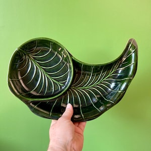 May include: A green ceramic serving dish shaped like a leaf with a smaller bowl attached. The dish has a raised design of veins and a glossy finish.