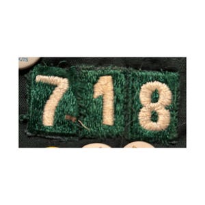 May include: A green embroidered patch with the numbers 718 in off-white thread. The numbers are stitched onto a dark green background. The patch is rectangular and has a textured appearance.