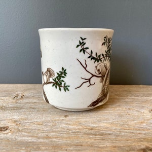 May include: White ceramic mug with a brown and green floral design featuring birds. The mug has a speckled glaze.