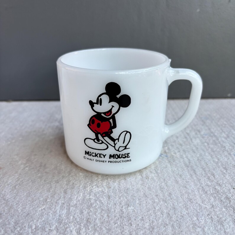 May include: White ceramic mug featuring a classic Mickey Mouse graphic in black, red, and white. The mug has a curved handle and the text "Mickey Mouse Walt Disney Productions" at the base. A vintage collectible.