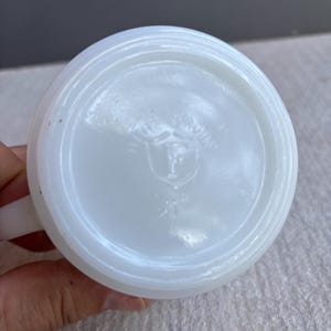 May include: A white, opaque glass mug with a handle. The bottom of the mug features a circular design with a shield and the letter "F" inside. The mug is clean and appears to be in good condition.