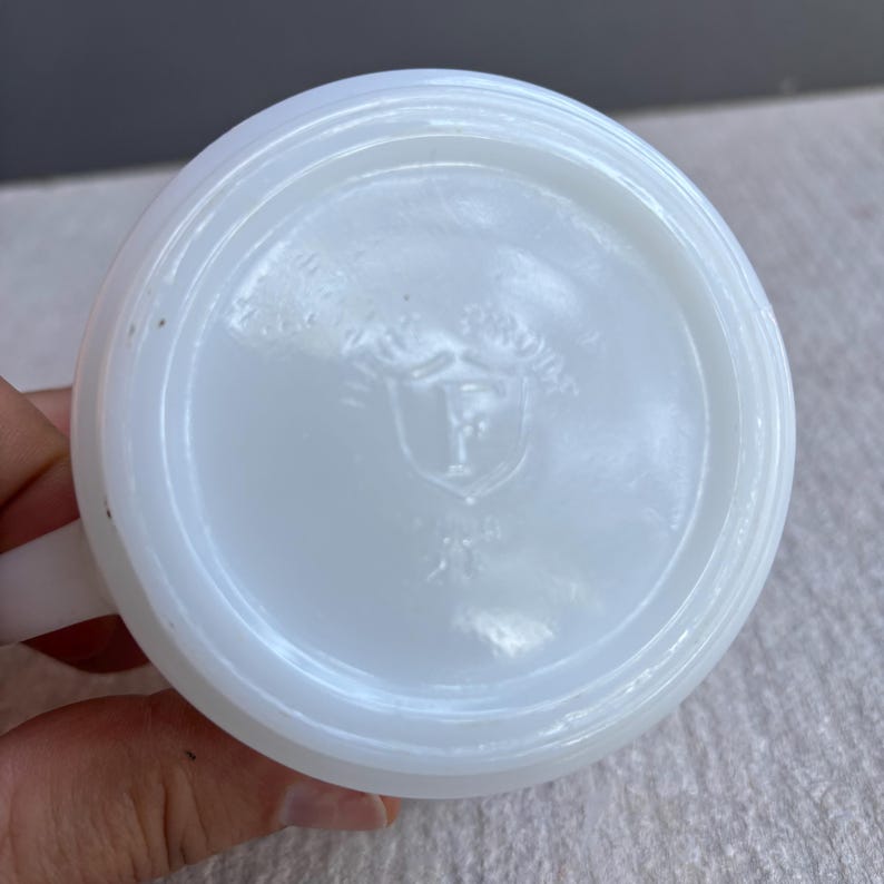 May include: A white, opaque glass mug with a handle. The bottom of the mug features a circular design with a shield and the letter "F" inside. The mug is clean and appears to be in good condition.